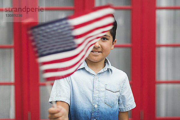 Porträt eines Jungen  der die amerikanische Flagge hält  während er gegen ein Haus steht