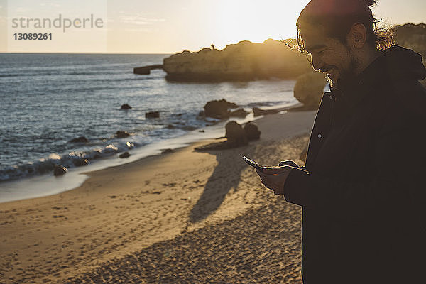 Lächelnder Mann benutzt Mobiltelefon  während er bei Sonnenuntergang am Strand gegen den Himmel steht