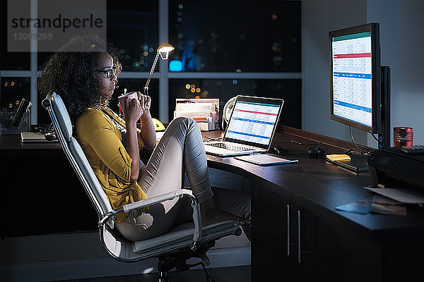 Seitenansicht einer nachdenklichen Geschäftsfrau  die eine Kaffeetasse hält  während sie abends im Büro auf einem Stuhl sitzt