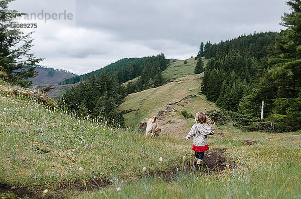 Rückansicht eines Mädchens  das auf einem Grasfeld mit einem Hund gegen Hügel läuft