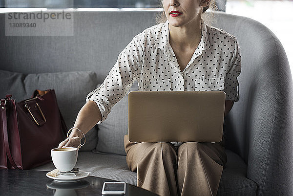 Geschäftsfrau hält Kaffeetasse bei der Arbeit am Laptop im Restaurant
