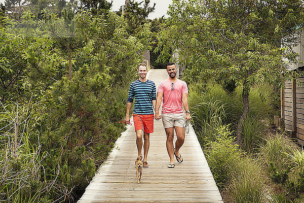 Homosexuelles Paar mit Chihuahua in voller Länge auf einer Strandpromenade inmitten von Bäumen