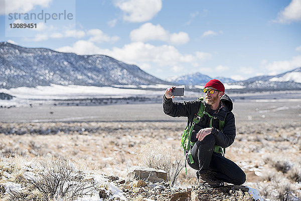 Wanderer fotografiert mit Smartphone in voller Länge im Winter in der Wüste gegen die Berge