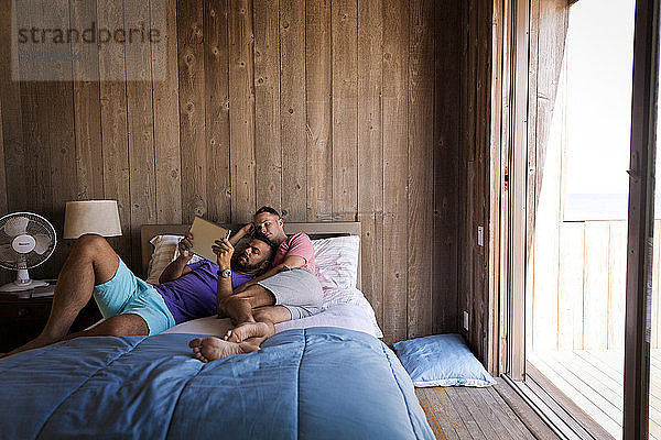 Schwule Männer benutzen einen Tablet-Computer  während sie auf dem Bett an einer Holzwand im Ferienort liegen