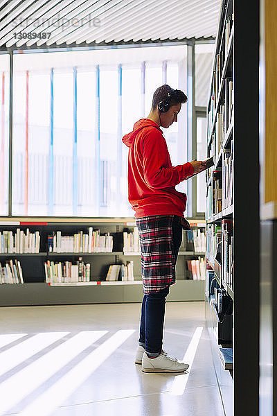 Seitenansicht eines Mannes mit Kopfhörern beim Lesen eines Buches im Stehen in der Bibliothek