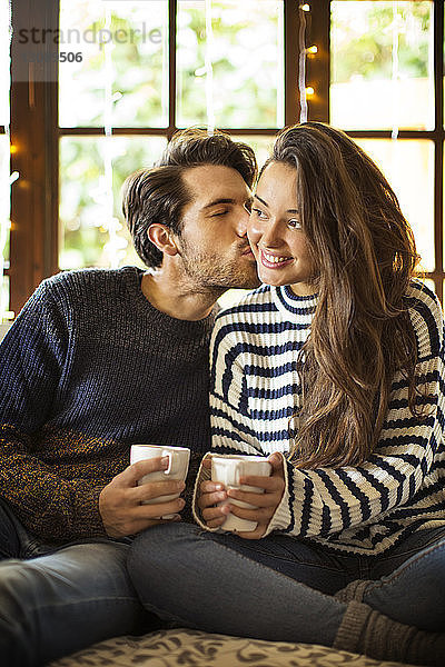 Mann küsst Freundin beim Kaffee trinken auf einem Fensterplatz in einer Nische zu Hause
