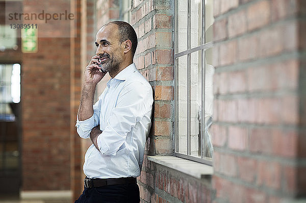 Seitenansicht eines Geschäftsmannes  der mit einem Smartphone spricht  während er an einer Ziegelmauer steht