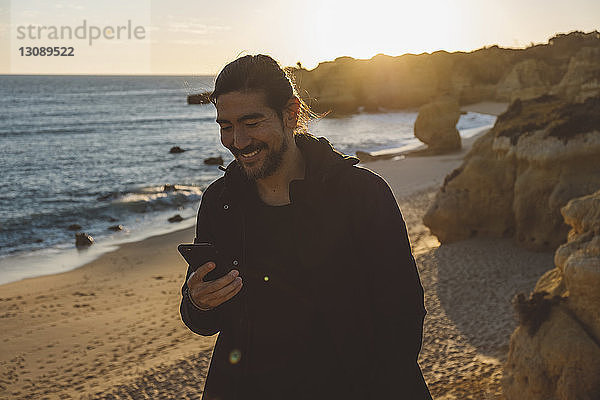 Lächelnder Mann benutzt Mobiltelefon  während er bei Sonnenuntergang am Strand steht