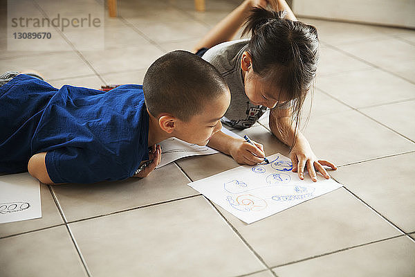 Bruder sieht Schwester an  die auf Papier zeichnet  während sie im Wohnzimmer auf dem Boden liegt