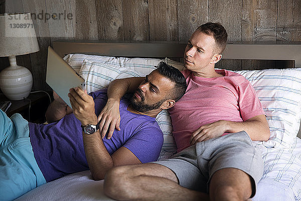 Homosexuelles Paar schaut auf Tablet-Computer  während es in einem Ferienort auf dem Bett liegt