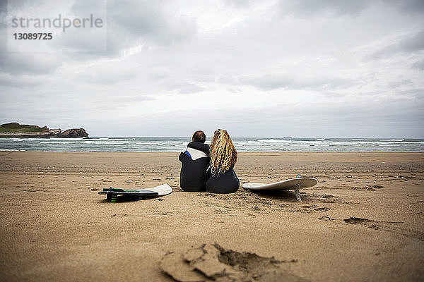 Rückansicht eines Paares  das mit Surfbrettern am Strand gegen den Himmel sitzt