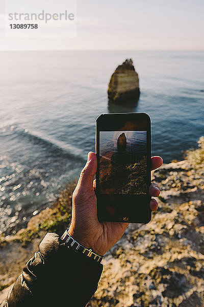 Abgetrennte Hand eines Mannes  der bei Sonnenuntergang das Meer mit einem Smartphone fotografiert