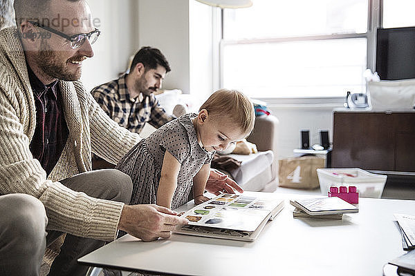 Vater zeigt der Tochter ein Bilderbuch  während der Partner im Hintergrund sitzt