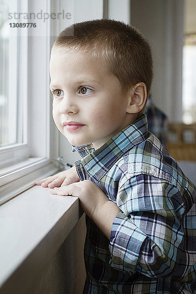 Lächelnder Junge schaut durchs Fenster  während er zu Hause steht