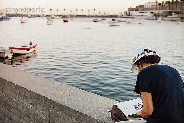 Hochwinkelansicht einer Frau  die malt  während sie auf der Stützmauer des Hafens sitzt