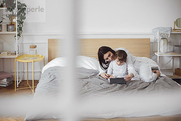 Glückliche Mutter  die einen Tablet-Computer benutzt  während der kleine Junge zu Hause im Bett liegt