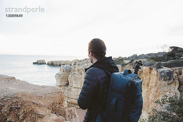 Seitenansicht eines Mannes mit Rucksack  der auf das Meer schaut  während er auf einer Klippe vor klarem Himmel steht