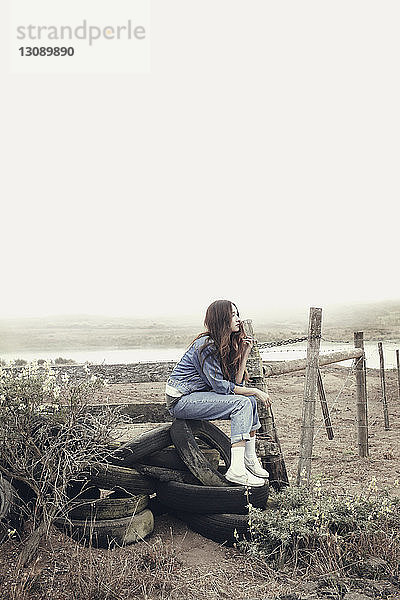 Nachdenkliche Frau sitzt auf verlassenen Reifen im Feld gegen den klaren Himmel