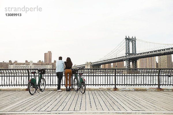Paar steht mit Fahrrädern auf Fußweg an der Manhattan-Brücke gegen den Himmel