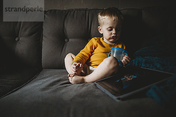 Süßer Junge mit Becher sitzt zu Hause auf dem Sofa