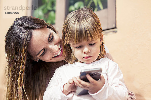 Glückliche Frau sieht ein Mädchen an  das im Haus ein Smartphone benutzt