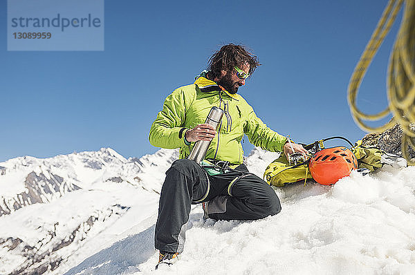 Männlicher Wanderer mit isoliertem Getränkebehälter sitzt auf schneebedecktem Berg vor strahlend blauem Himmel
