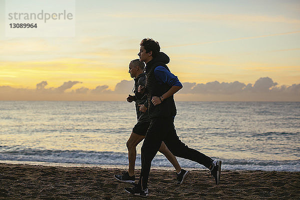 Entschlossener Vater und Sohn joggen am Strand vor bewölktem Himmel bei Sonnenuntergang