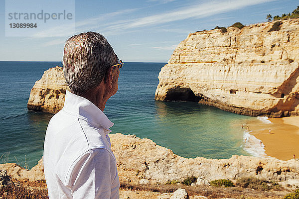 Älterer Mann schaut aufs Meer  während er auf einer Klippe gegen den Himmel steht