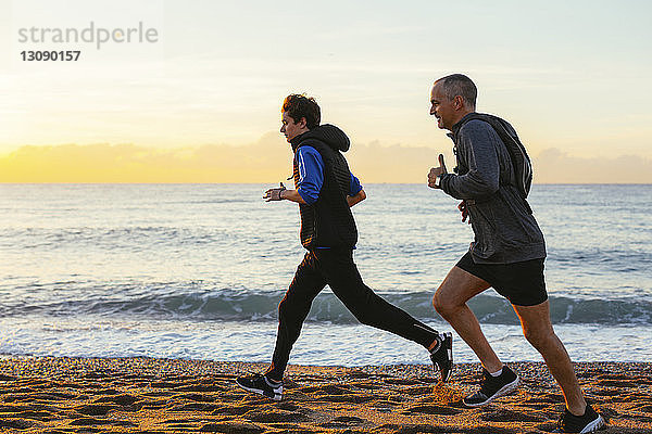 Vater und Sohn joggen in voller Länge an der Küste am Strand gegen den Himmel während des Sonnenuntergangs