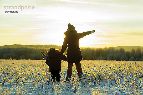 Rückansicht von Mutter und Sohn  die bei Sonnenuntergang auf einem verschneiten Feld gegen den Himmel stehen