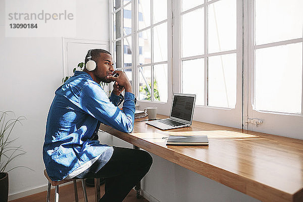 Seitenansicht eines nachdenklichen Mannes  der Musik hört  während er mit einem Laptop-Computer an einem Holztisch am Fenster sitzt