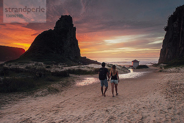 Rückansicht eines Paares  das bei Sonnenuntergang auf Sand gegen den Himmel läuft