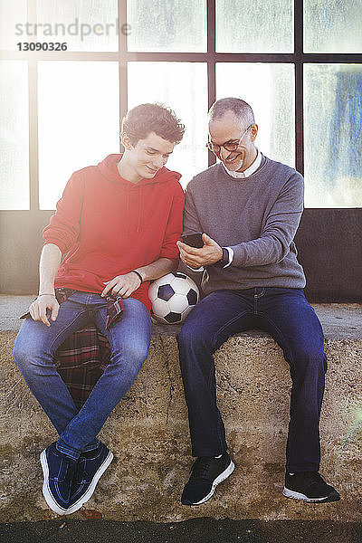 Glücklicher Vater zeigt seinem Sohn ein Smartphone  während er auf einer Stützmauer sitzt