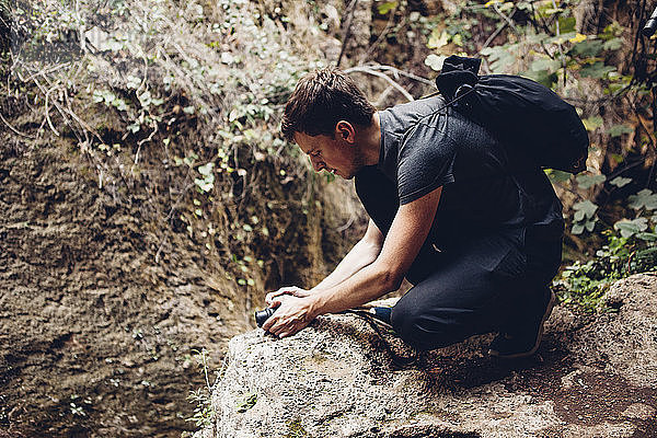 Männlicher Wanderer mit Rucksack fotografiert durch die Kamera  während er auf einer Felsformation kauert