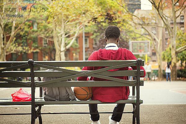 Rückansicht eines Schülers  der auf einer Bank am Basketballplatz sitzt