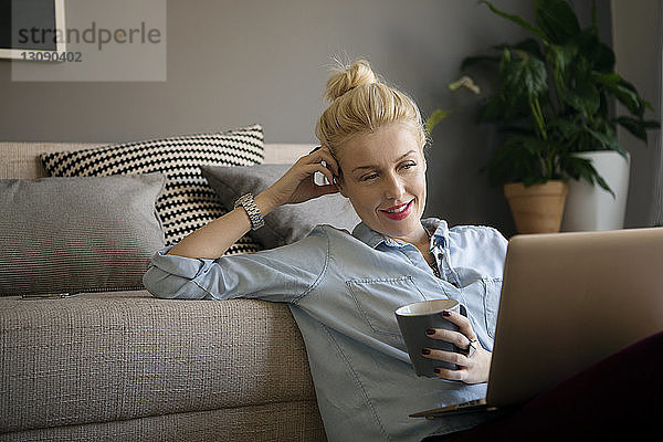 Lächelnde Frau hält Kaffeetasse  während sie im Wohnzimmer einen Laptop benutzt