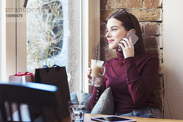 Frau mit Getränk schaut durchs Fenster  während sie im Cafe mit dem Handy telefoniert