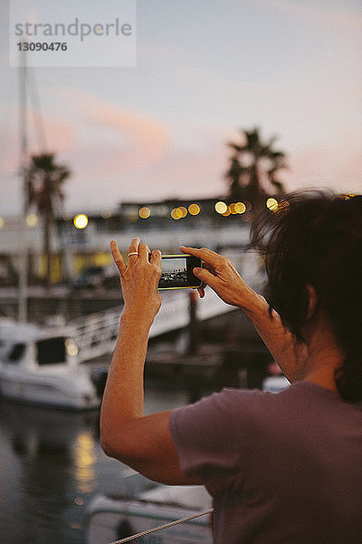Rückansicht einer Frau  die bei Sonnenuntergang am Hafen mit einem Smartphone fotografiert