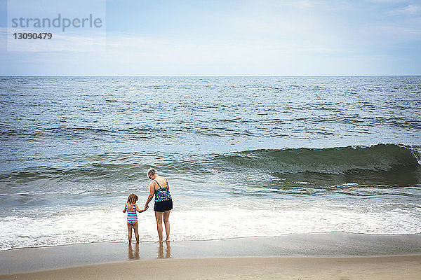 Rückansicht der Enkelin und Großmutter  die am Strand am Ufer stehen und Händchen halten