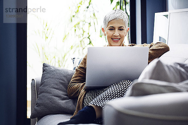 Lächelnde Frau benutzt Laptop  während sie zu Hause auf dem Sofa sitzt