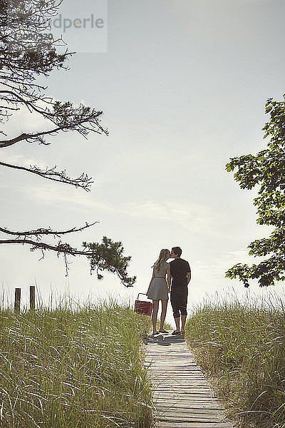 Rückansicht eines Paares  das sich küsst  während es auf einer Strandpromenade inmitten eines Feldes gegen den Himmel steht