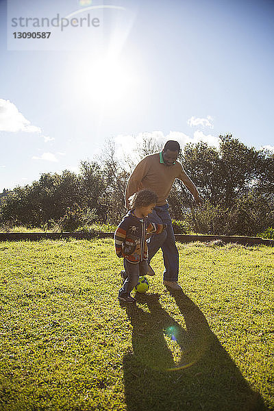 Vater und Sohn spielen bei Sonnenschein mit dem Ball auf einem Grasfeld gegen den Himmel