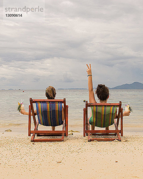 Rückansicht von Freundinnen  die sich auf Liegestühlen am Strand vor bewölktem Himmel entspannen