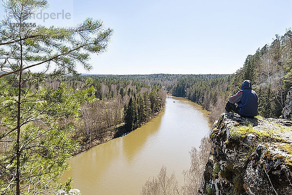 Rückansicht eines Mannes  der eine schöne Aussicht hat  während er auf einer Klippe vor klarem  blauem Himmel sitzt