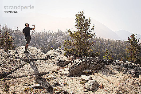 Rückansicht eines Mannes  der auf einem Fels steht und an einem sonnigen Tag gegen Berge fotografiert
