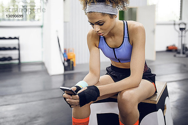 Sportlerin benutzt Smartphone  während sie im Fitnessstudio auf der Box sitzt