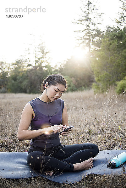 Frau hört Musik  während sie ein Smartphone auf der Trainingsmatte auf dem Feld benutzt