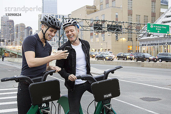 Fröhliche Freunde schauen auf ihr Handy  während sie mit Fahrrädern auf der Straße der Stadt stehen