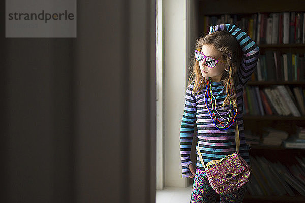 Mädchen mit Halskette und Sonnenbrille steht zu Hause am Fenster