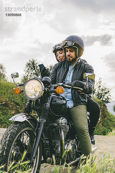 Ehepaar benutzt Mobiltelefon  während es auf einem Motorrad gegen den Himmel sitzt